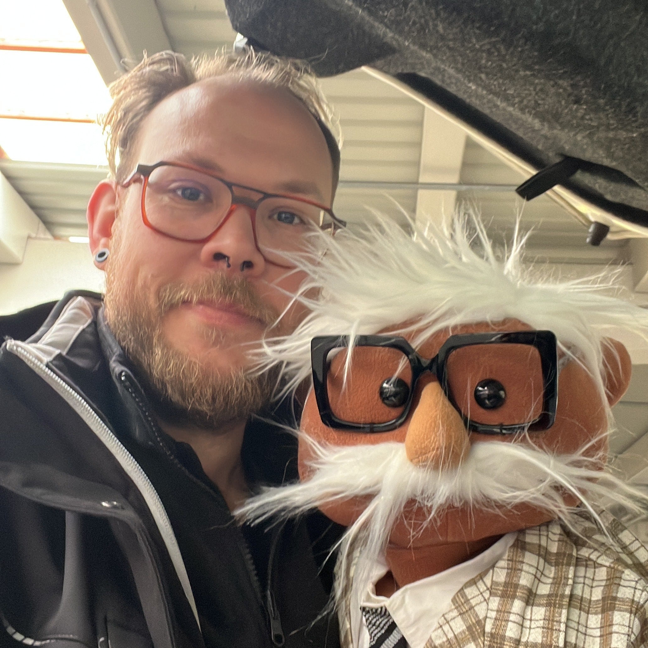 Person with a beard and glasses next to a puppet with similar features, inside a vehicle.