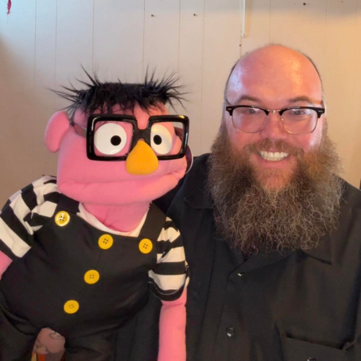 Person with a beard and glasses sitting next to a pink puppet character with glasses and a striped outfit.