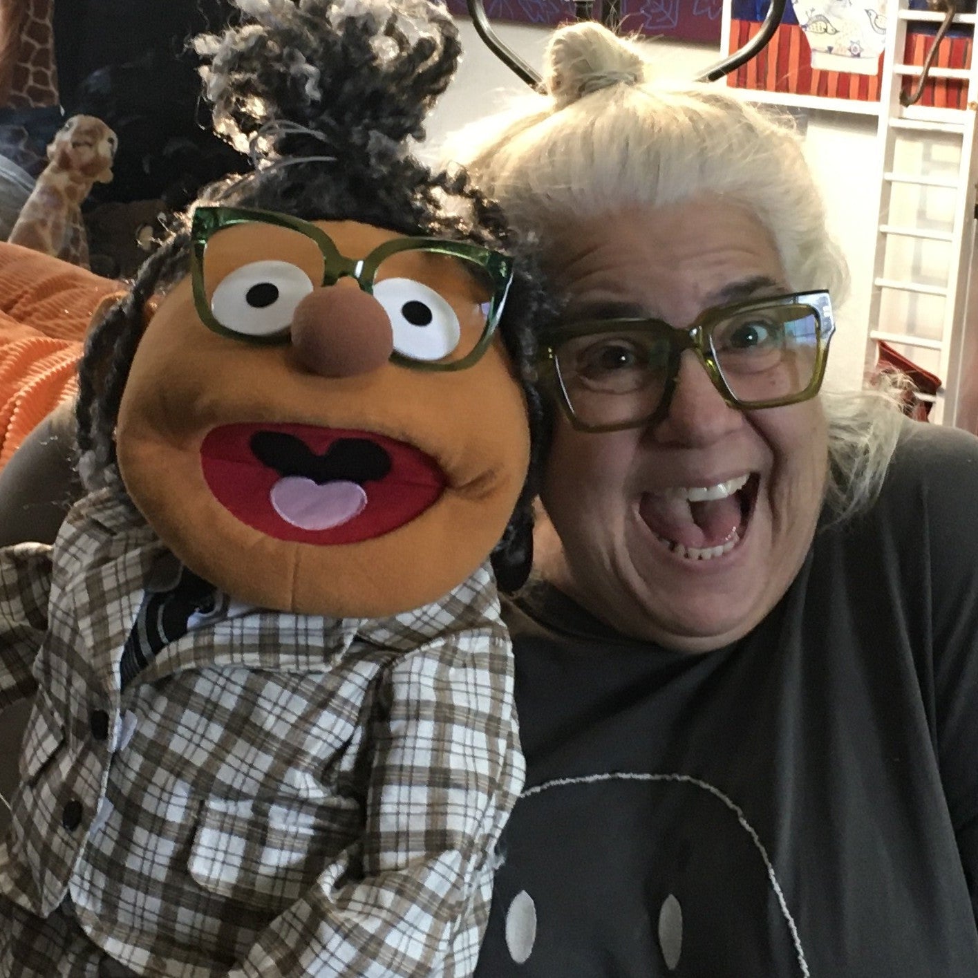 Person with a large plush puppet wearing glasses in a room with shelves and furniture.