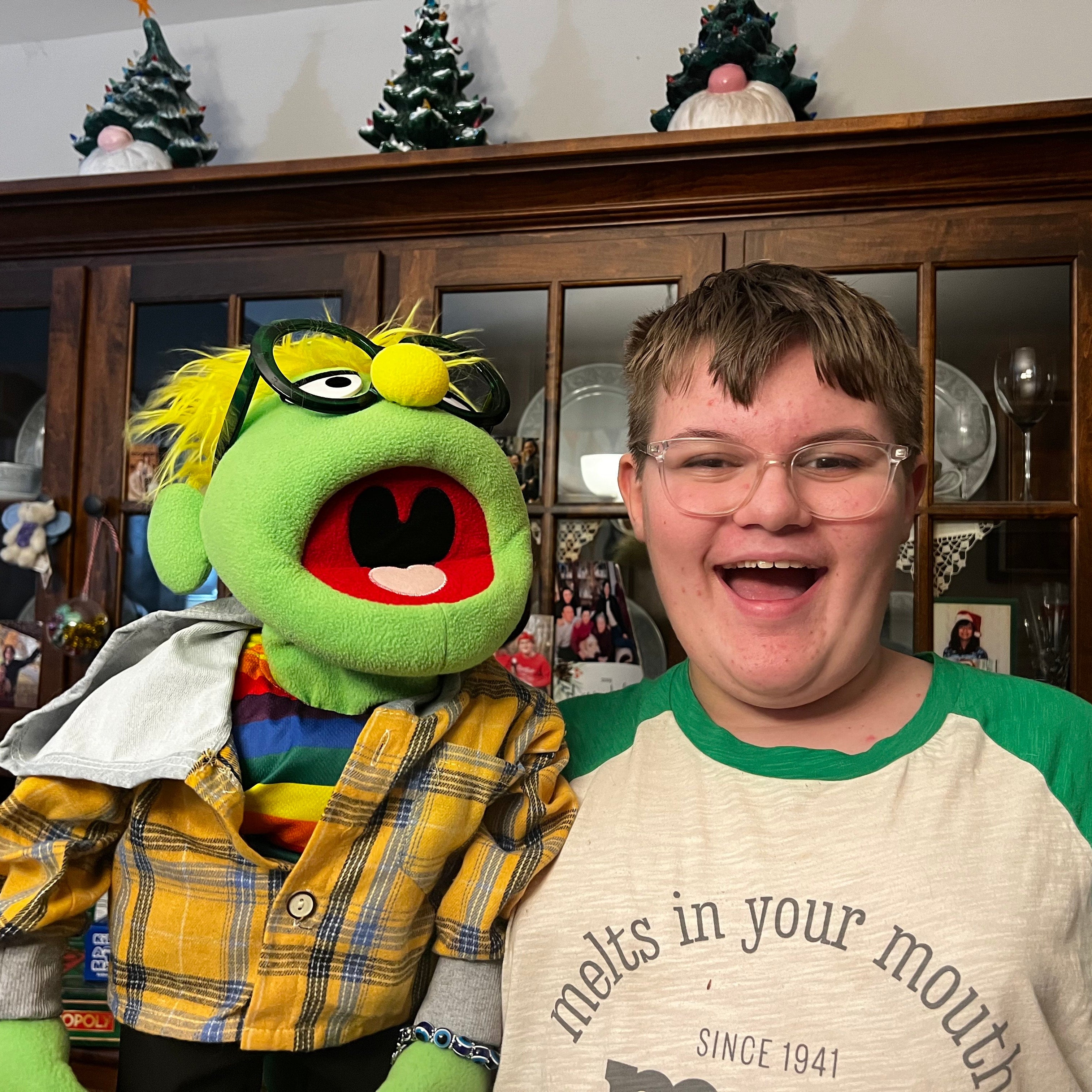 Person with a green puppet in a room with a wooden cabinet and decorative items.