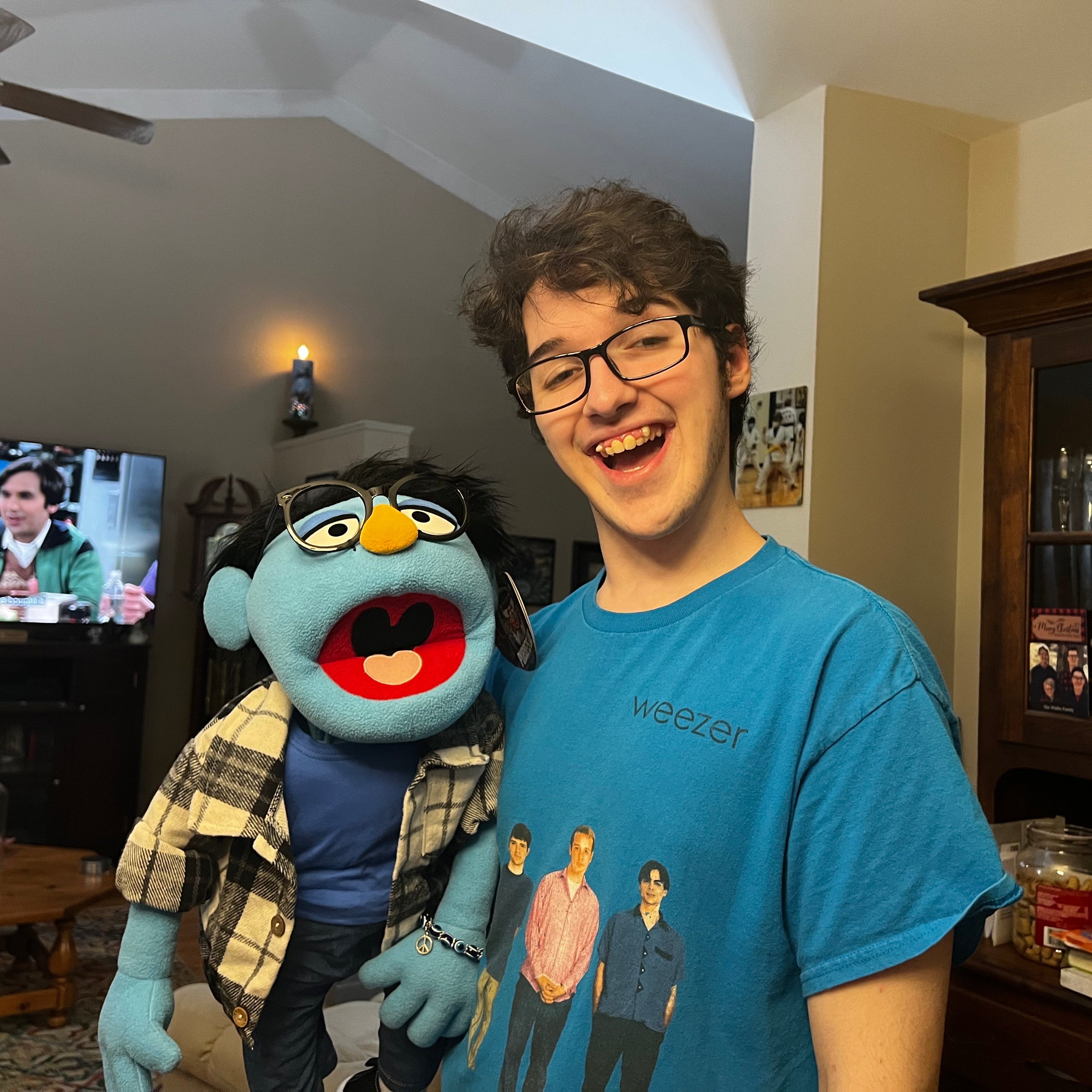 Person wearing a blue 'weezer' t-shirt with a puppet in a living room.