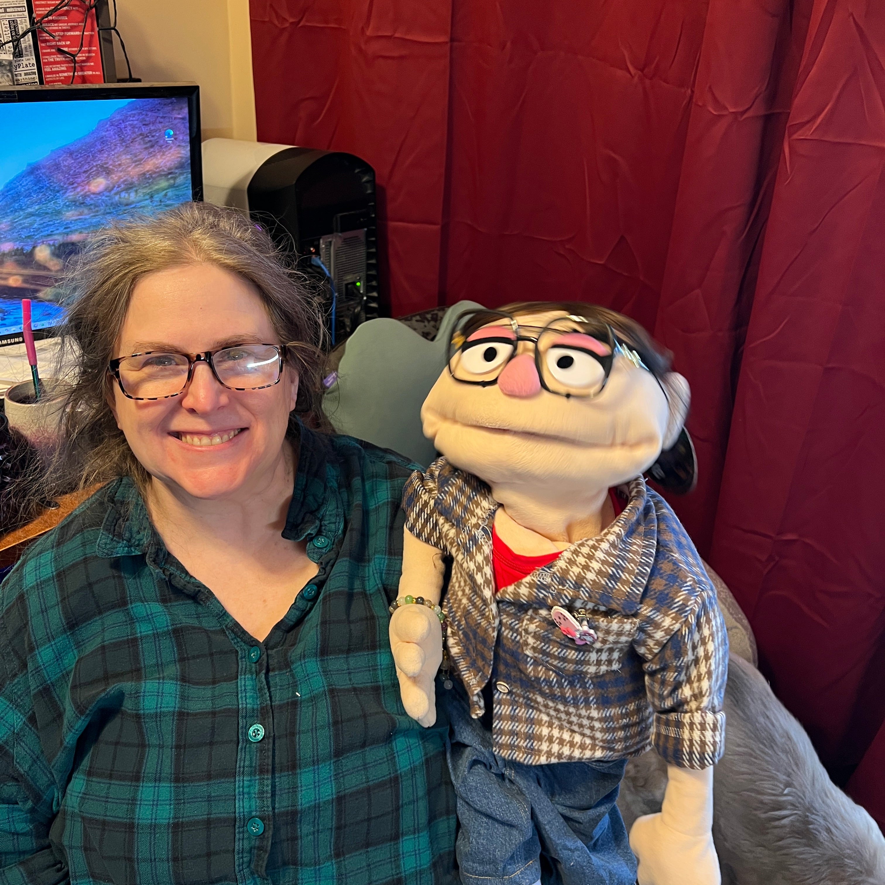 Person wearing a plaid shirt with a cartoonish puppet on their shoulder, in a room with a red curtain.
