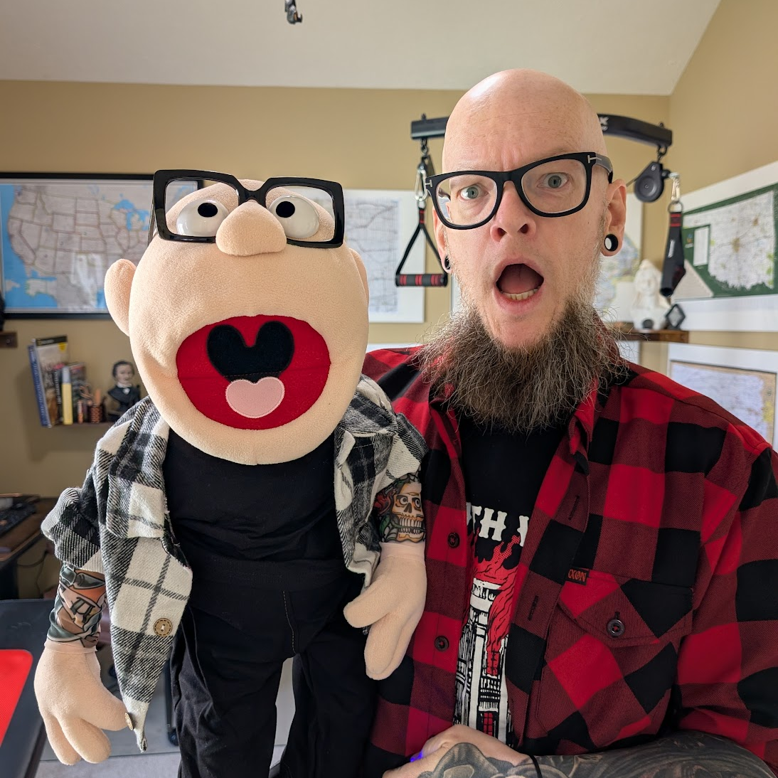 Person with tattoos and a red plaid shirt holding a puppet with glasses and a heart-shaped nose.