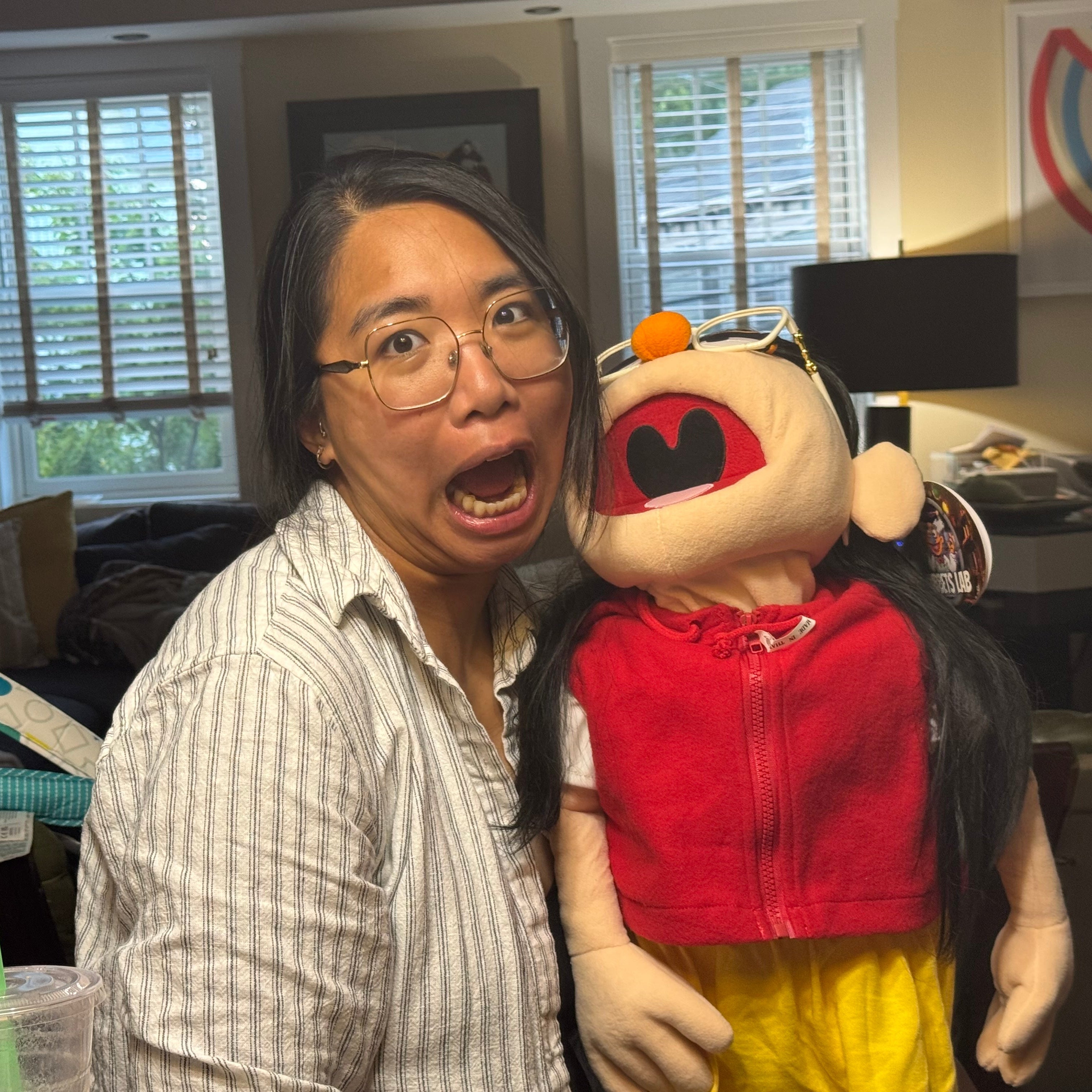 Person with glasses holding a puppet toy in a room with furniture and decor.